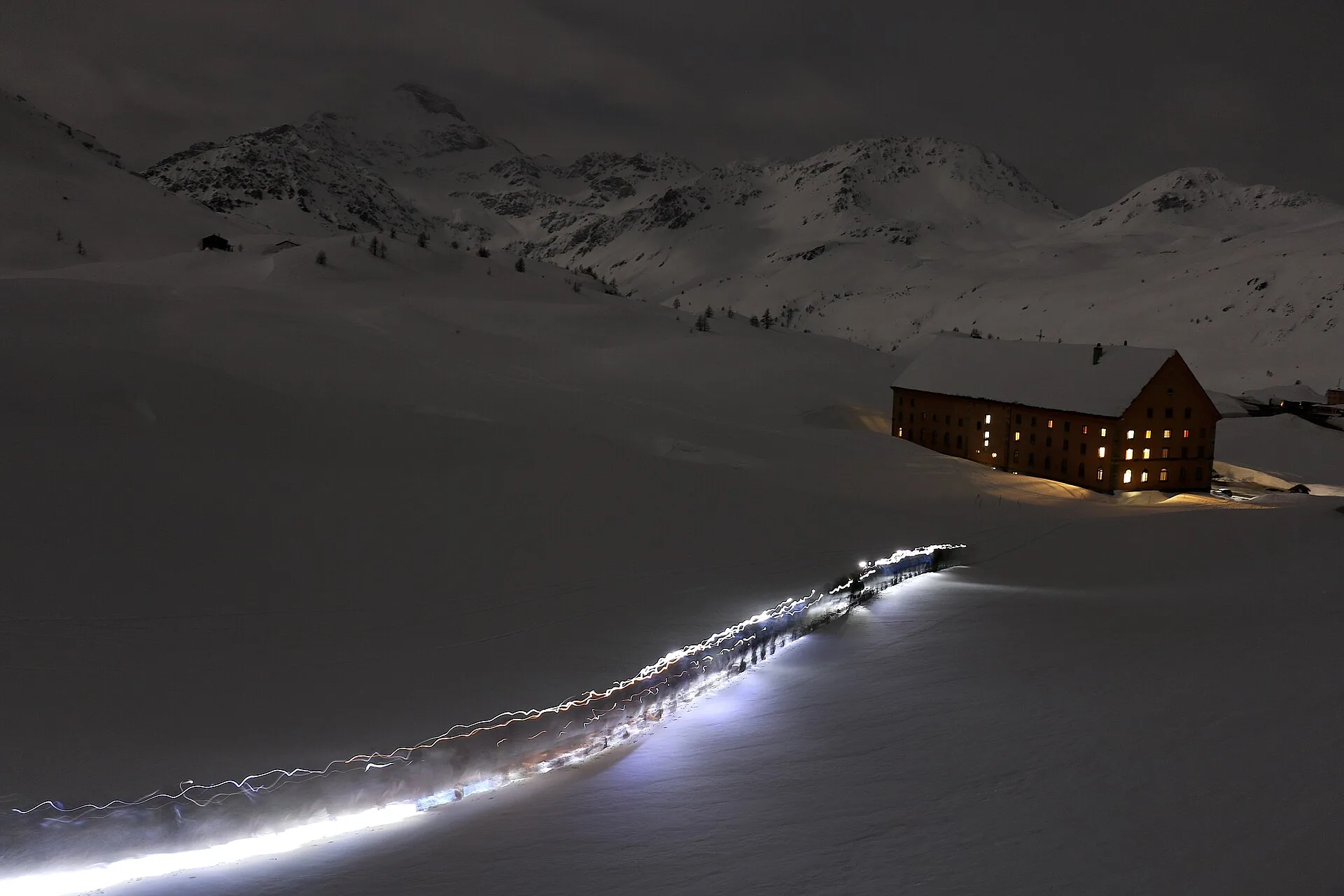 Escursionista con la frontale in marcia di sera verso un rifugio illuminato, sul passo del Sempione in inverno