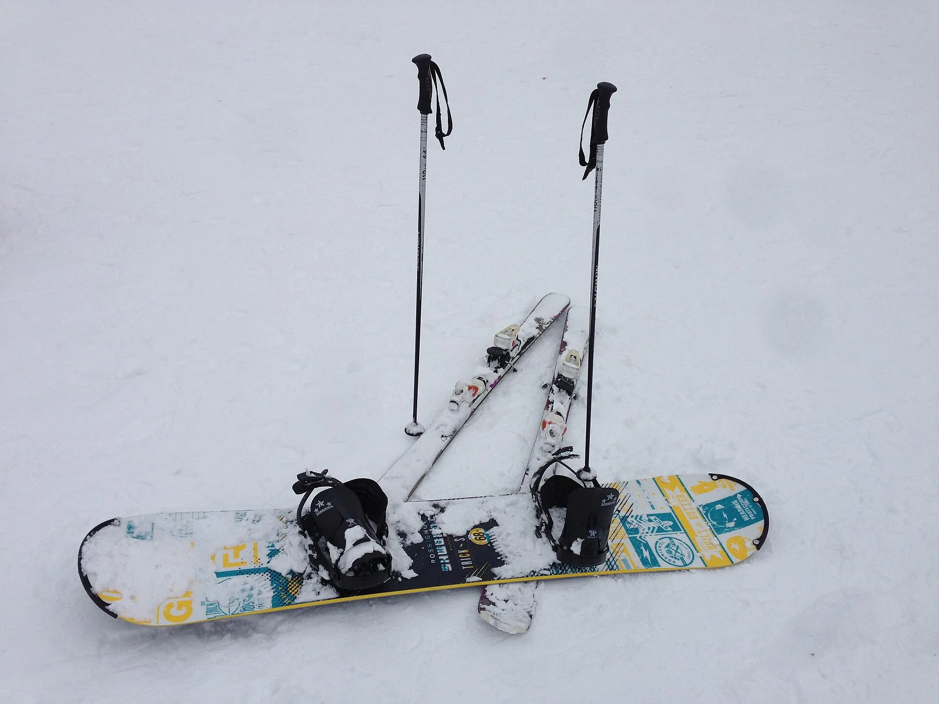 Snowboard con attacchi appoggiato sulla neve accanto a bastoncini e sci